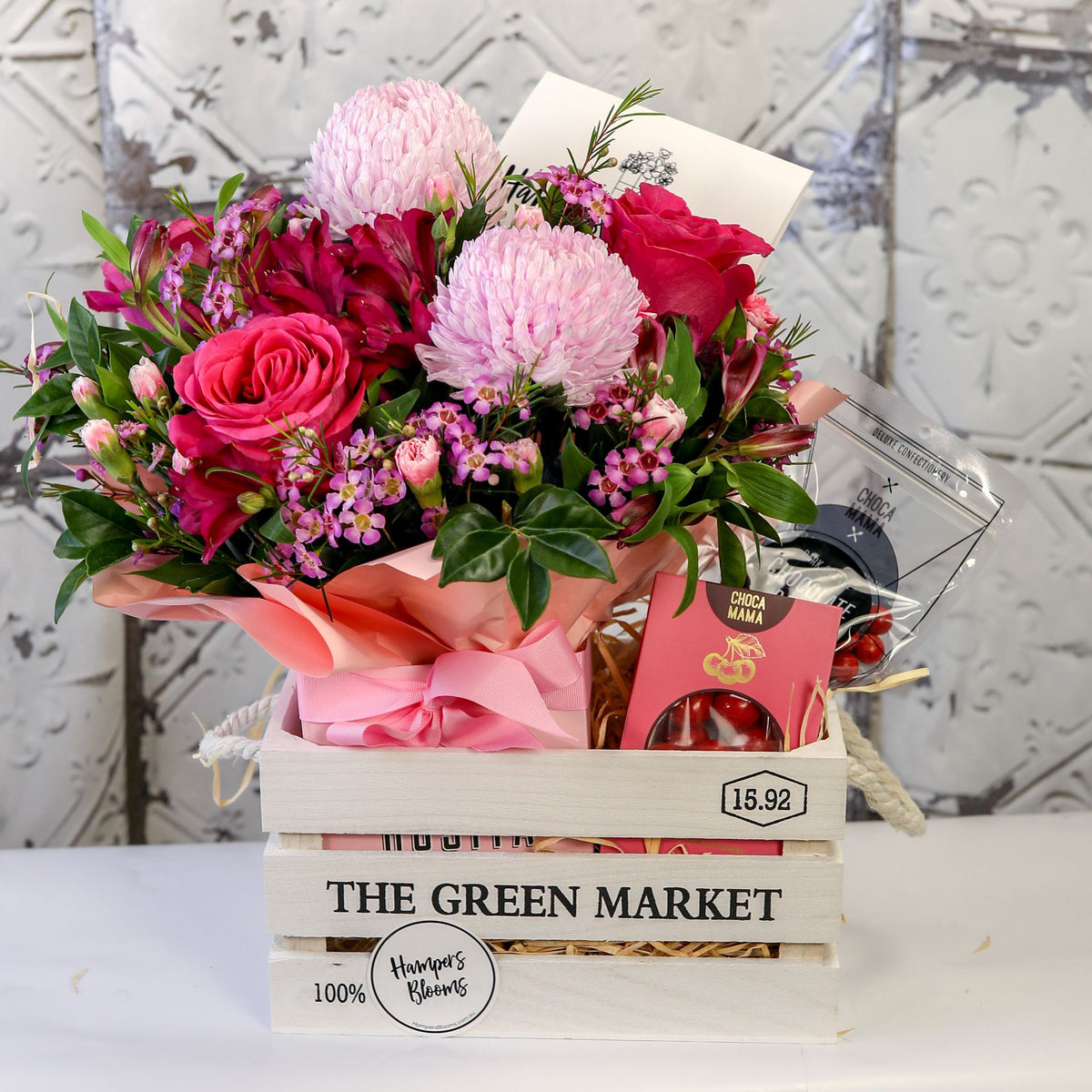Angled view of pink blooms in pink wrap and chocolates above the front of a white crate