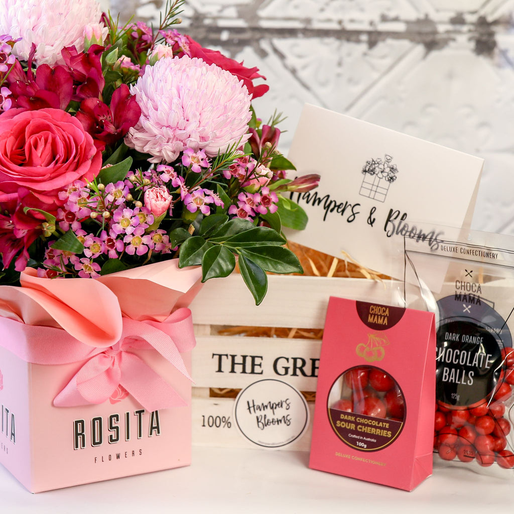 Close-up of pink flowers in a ribbon-wrapped pot beside a white crate, message card and chocolates
