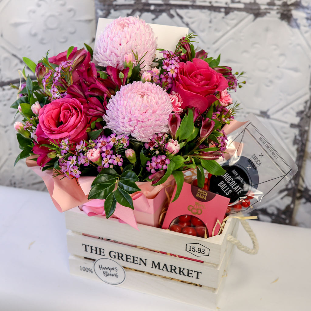 Angled view of pink flowers in a pink-wrapped pot and chocolates inside a white crate