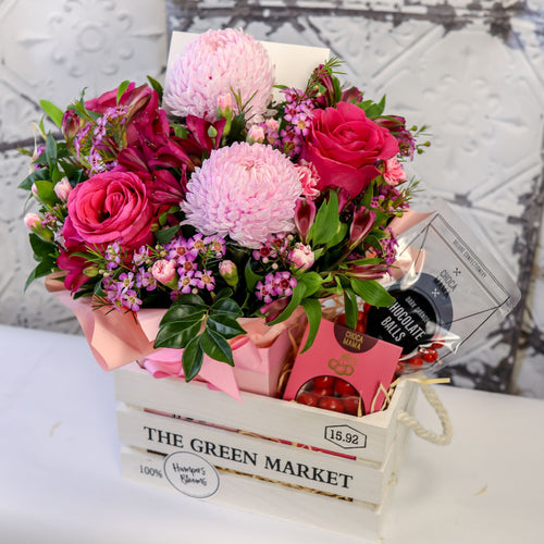 Angled view of pink flowers in a pink-wrapped pot and chocolates inside a white crate