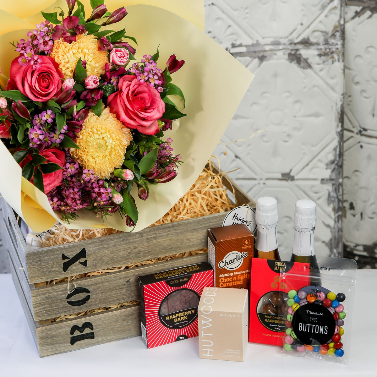Close-up of bright flowers over a timber crate with bottles, chocolates, biscuits and candle box displayed beside it