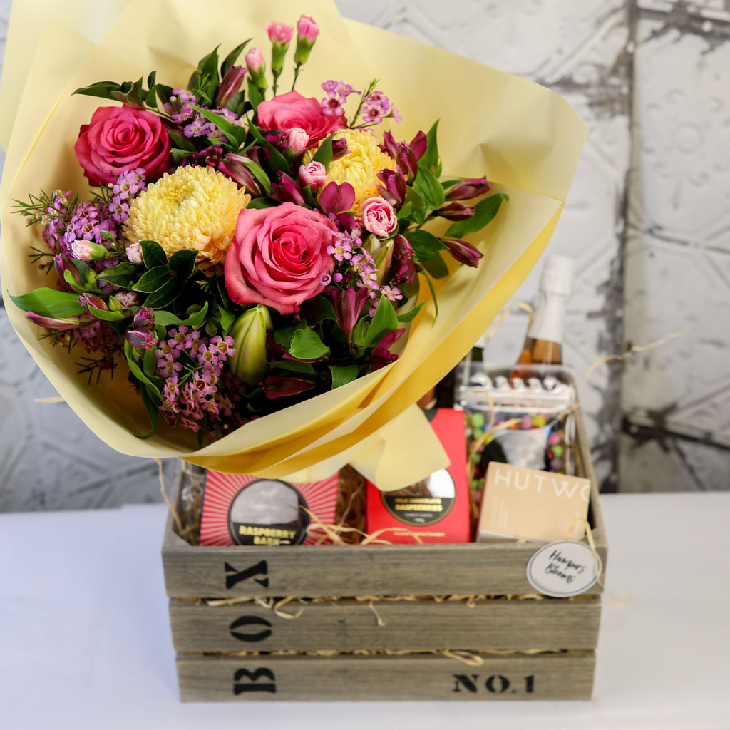 Front view of bright flowers in yellow wrap above chocolates and a candle box in a timber crate