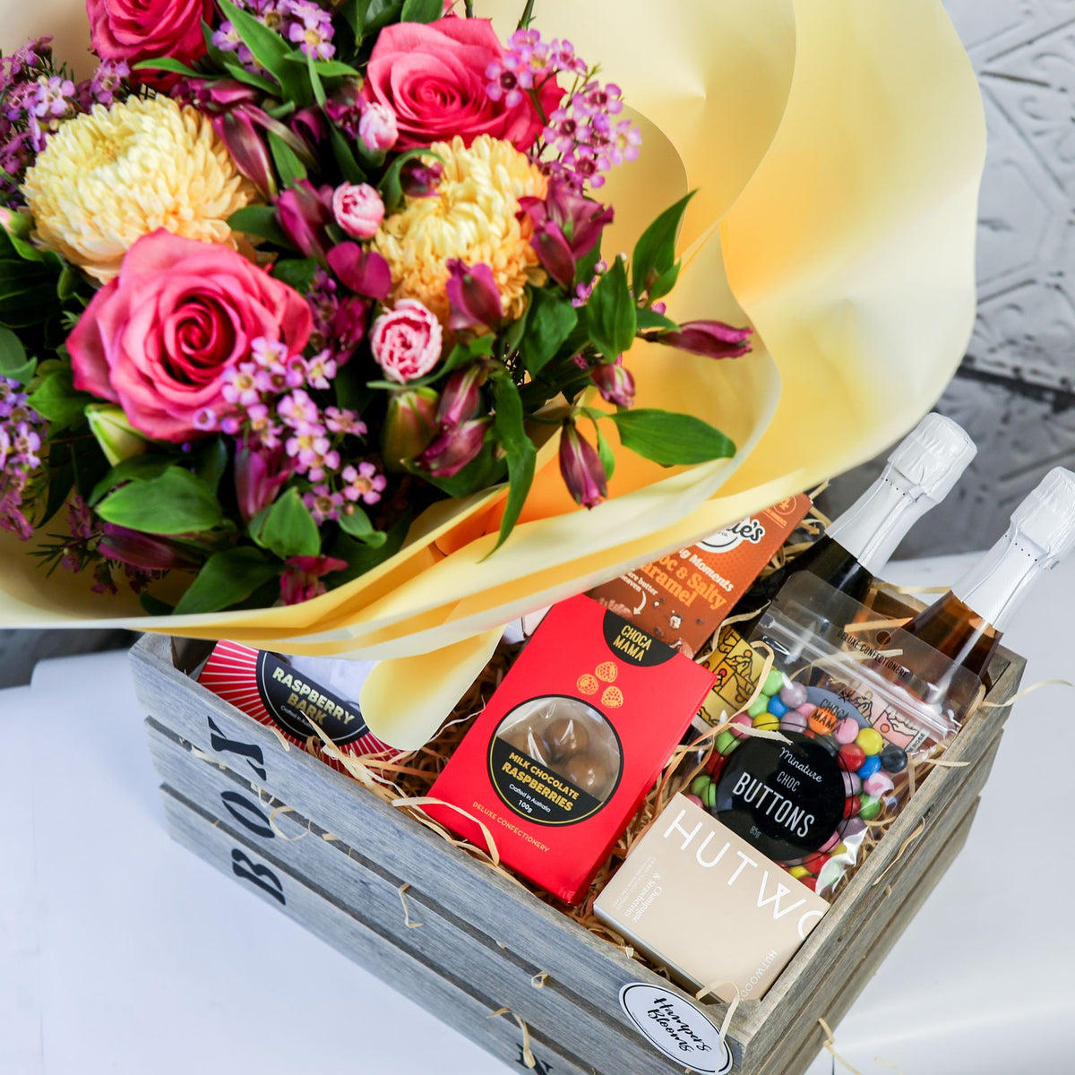 View from above of bright flowers, two small bottles, chocolates and candle box in a timber crate