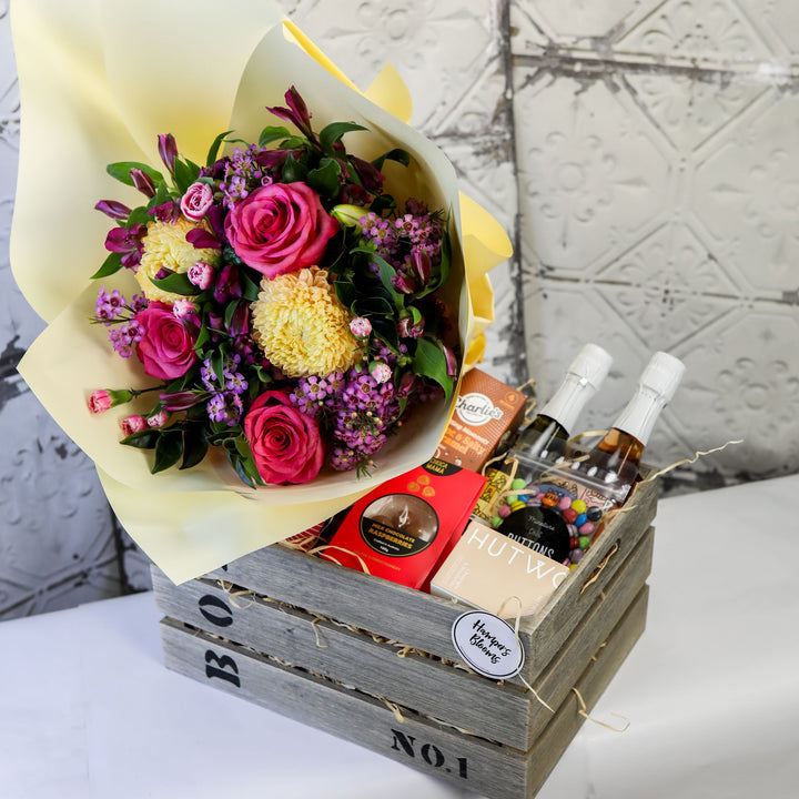 Bright pink and yellow bouquet, two small sparkling bottles and treats in a timber crate
