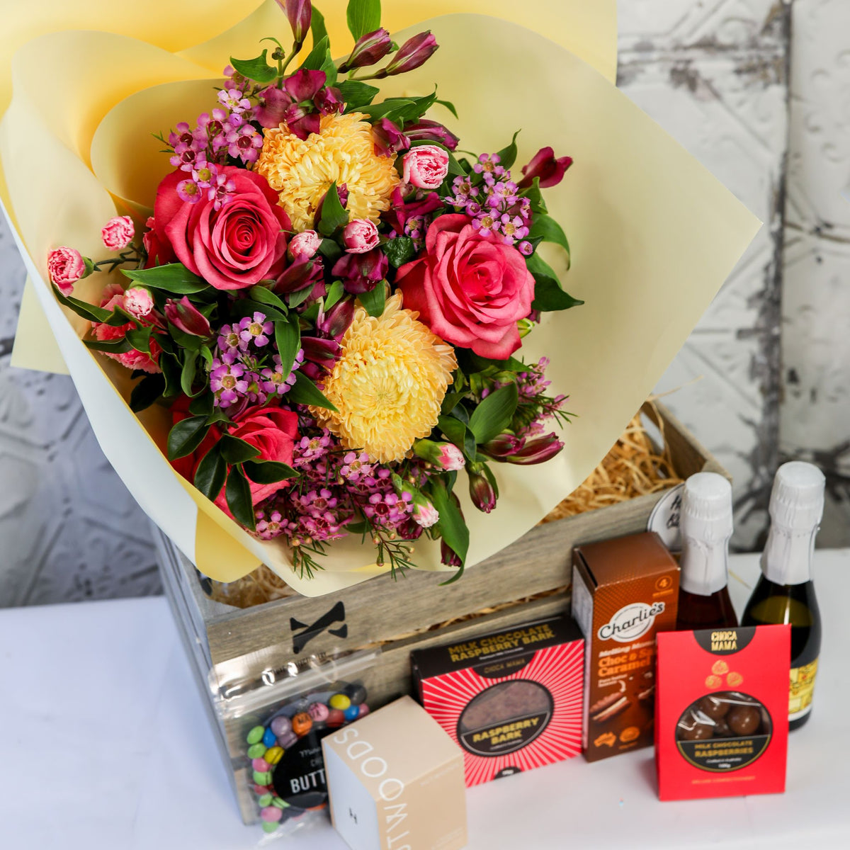 Bright bouquet over a timber crate with two small bottles, chocolates, biscuits and candle box displayed in front