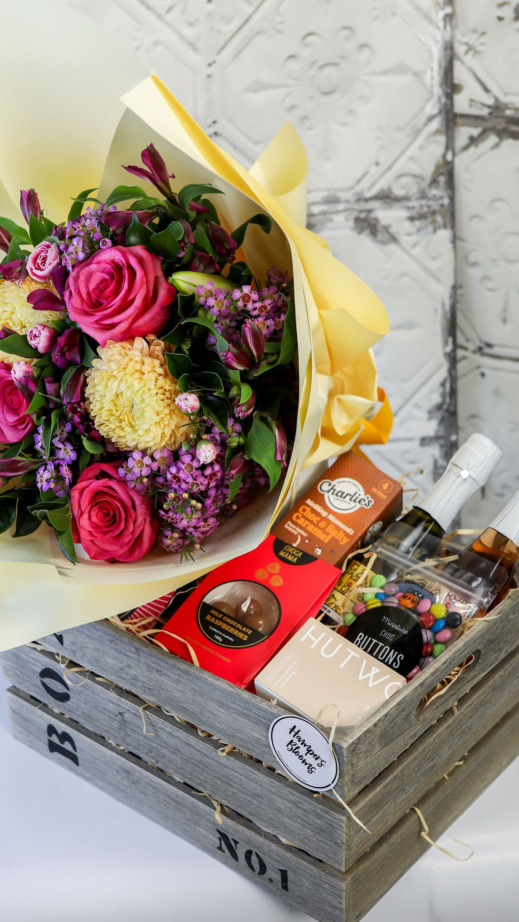 Portrait crop of bright flowers in yellow wrap above two small bottles and treats in a timber crate