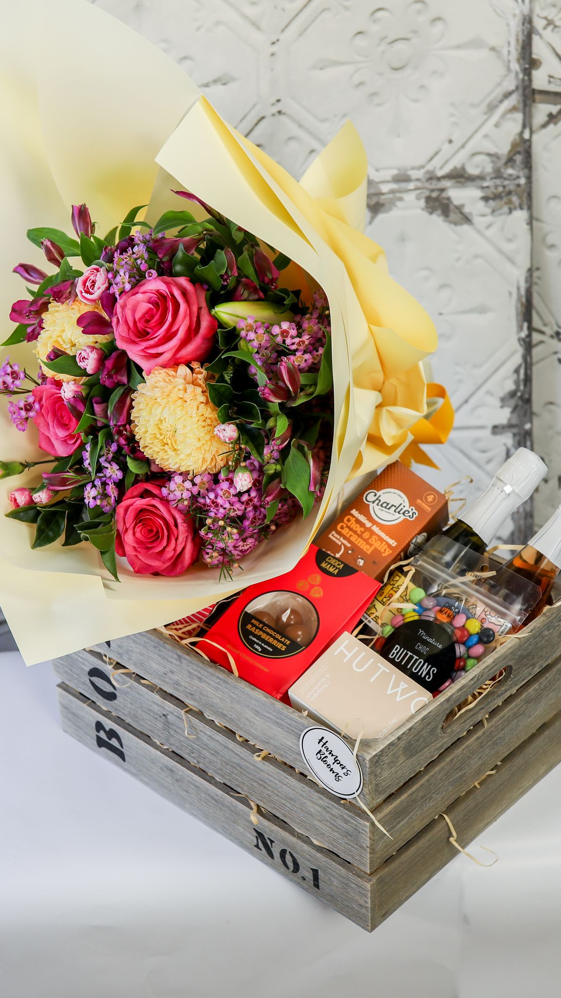 Angled view of a bright bouquet, two small bottles and treats arranged in a timber crate