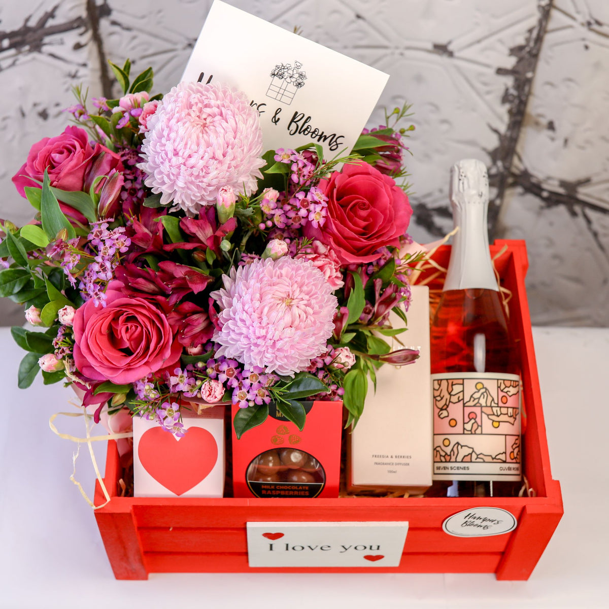 View from above of pink flowers, bottle, diffuser box and treats arranged inside a red crate