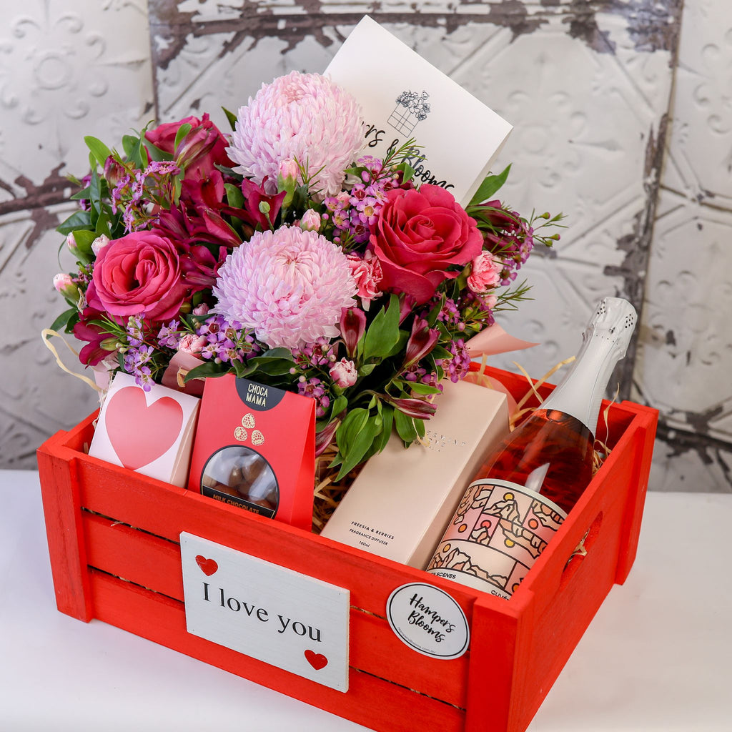 Angled view of pink blooms, bottle, diffuser box and treats arranged inside a red crate
