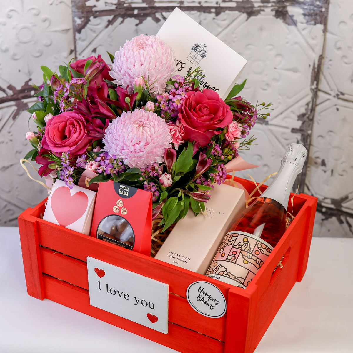 Angled view of pink blooms, bottle, diffuser box and treats arranged inside a red crate
