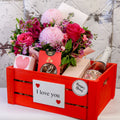 Pink floral arrangement, sparkling wine bottle and treat packages in a large red I love you crate