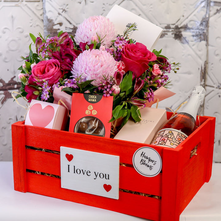 Pink floral arrangement, sparkling wine bottle and treat packages in a large red I love you crate