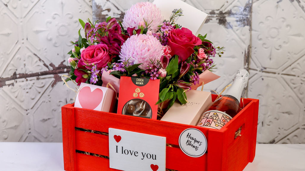 Landscape view of pink blooms, bottle and treats arranged in a red I love you crate