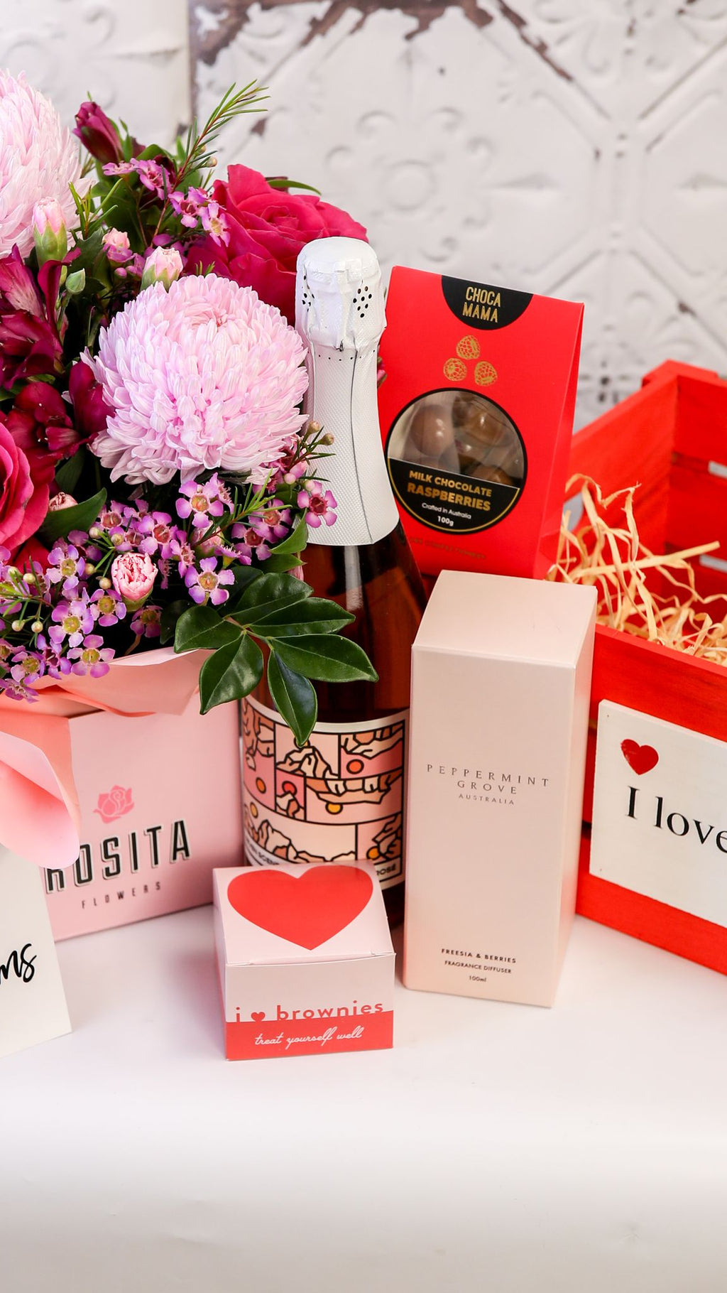 Portrait of pink flowers, bottle, diffuser box and treats displayed beside a red I love you crate