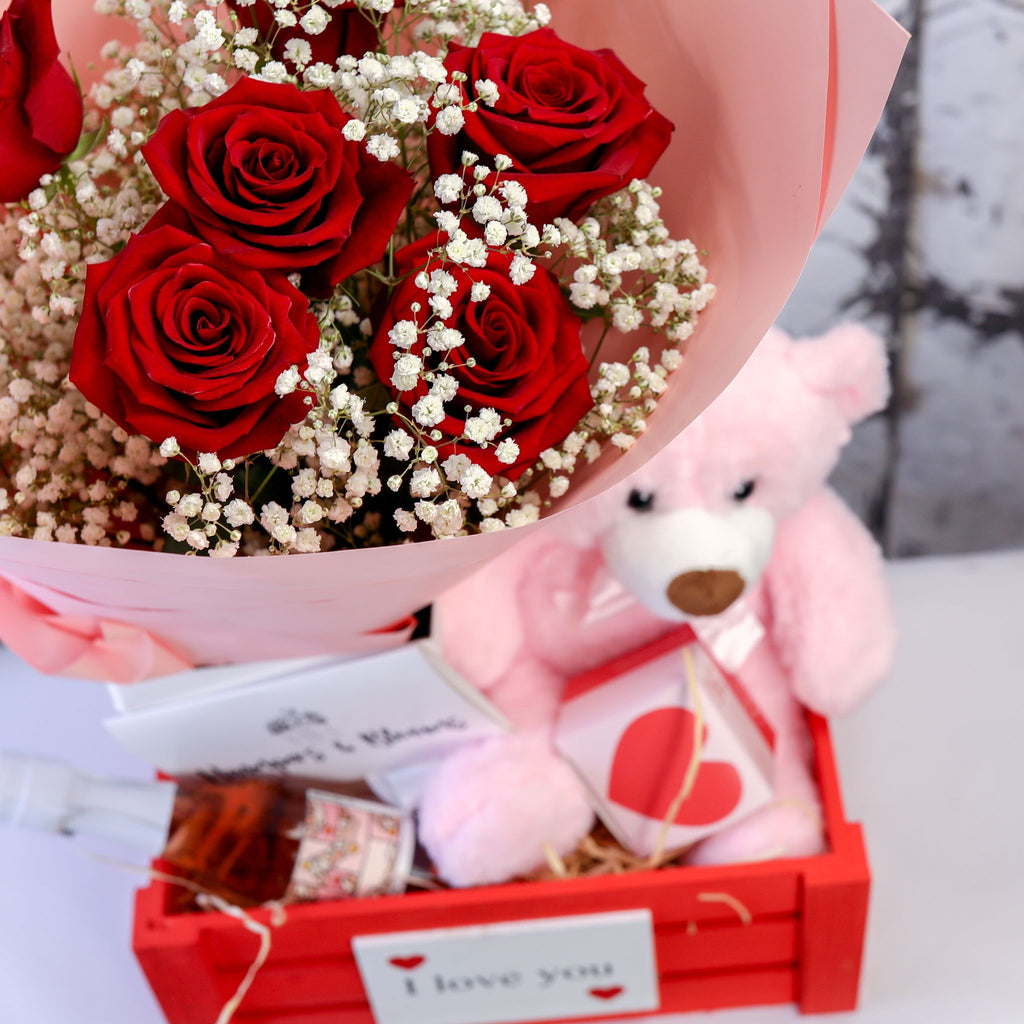 Close-up of red roses, pink teddy and treat packages arranged in a red crate