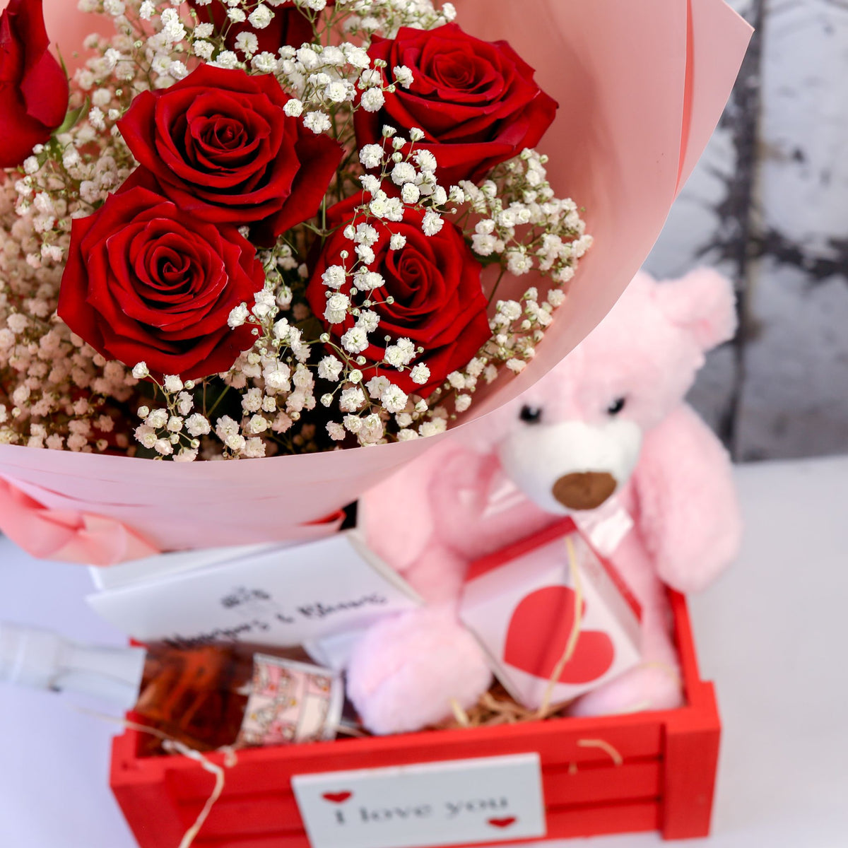 Close-up of red roses, pink teddy and treat packages arranged in a red crate