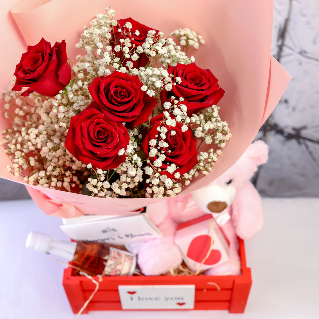 View from above of pink-wrapped red roses, pink teddy and treats in a red crate