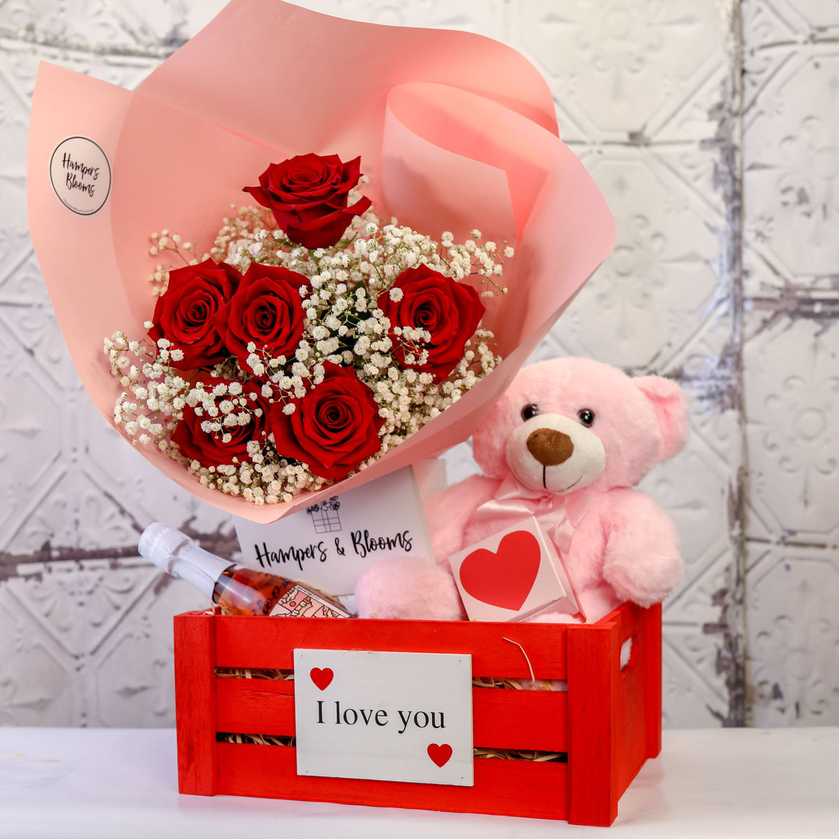 Front view of red roses in pink wrap and pink teddy above a red I love you crate