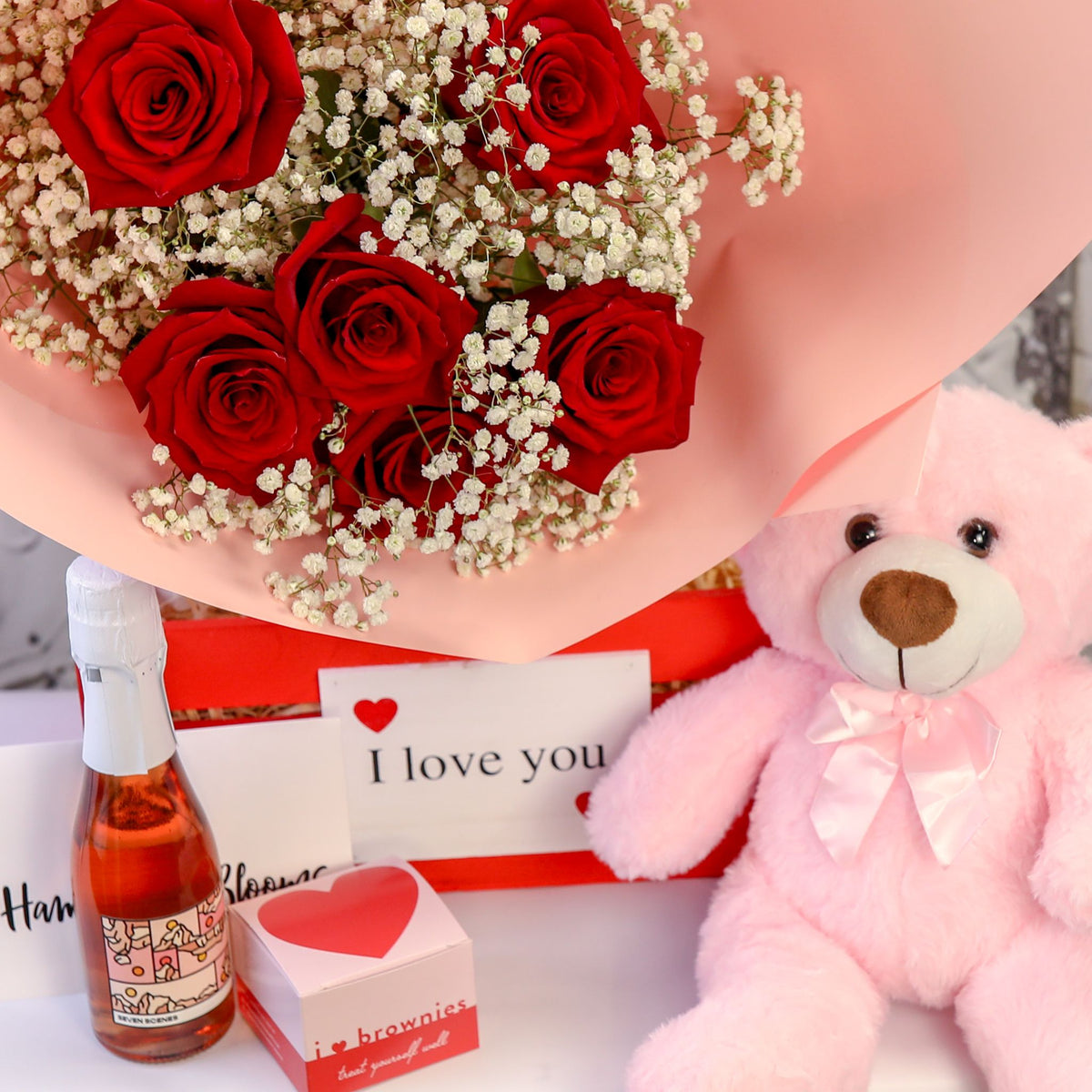 Close-up of red roses above a red crate with a pink teddy, small bottle and heart box displayed in front