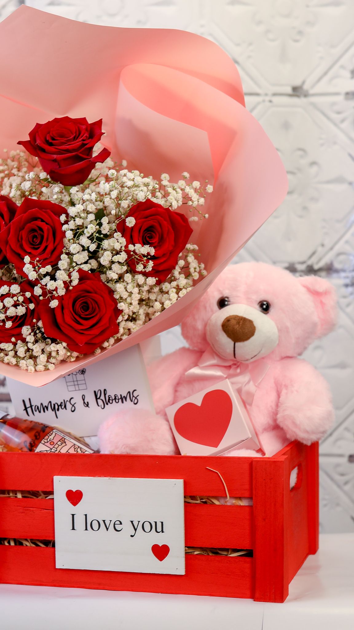 Portrait view of pink-wrapped red roses and pink teddy in a red I love you crate