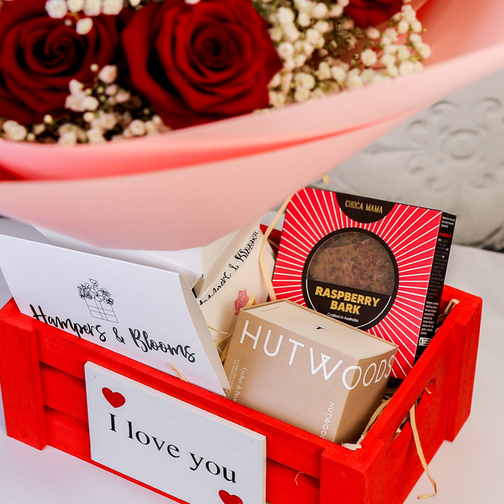Close-up of chocolates, boxed candle and message card in a red crate beneath pink-wrapped roses