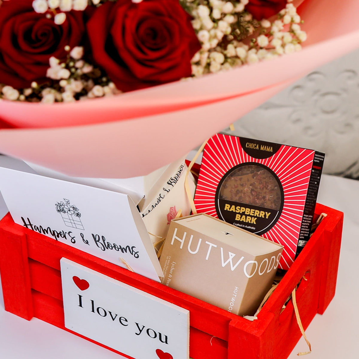 Close-up of chocolates, boxed candle and message card in a red crate beneath pink-wrapped roses