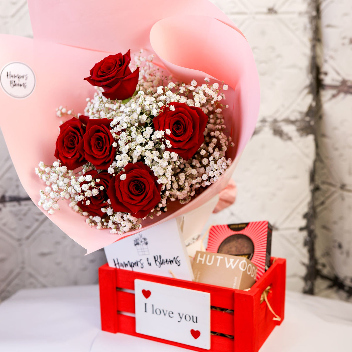 Angled view of red roses, chocolates, candle box and message card in a small red crate