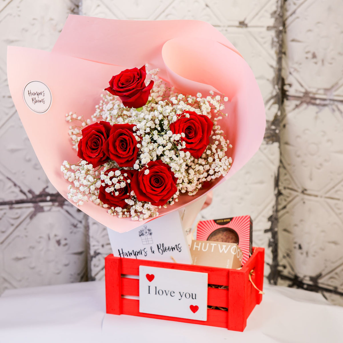 Red rose bouquet in pink wrap with chocolates and boxed candle in a small red I love you crate