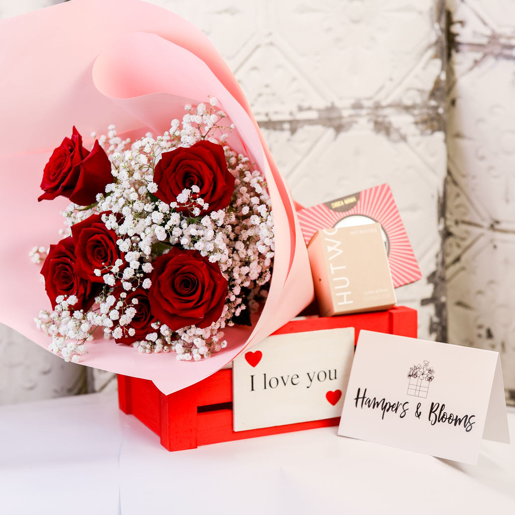 Red rose bouquet in a red I love you crate with candle and chocolate boxes displayed behind it and a card beside it