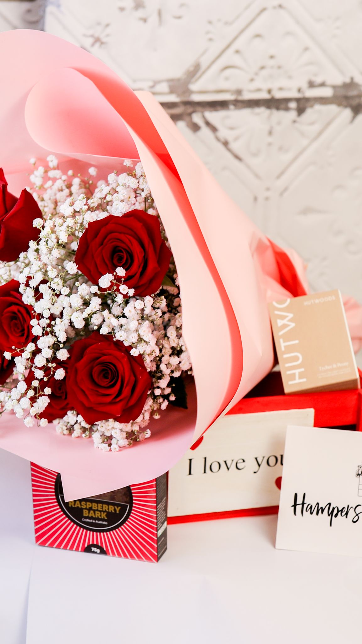 Portrait crop of pink-wrapped red roses and a red crate with chocolates, candle box and a card displayed around it