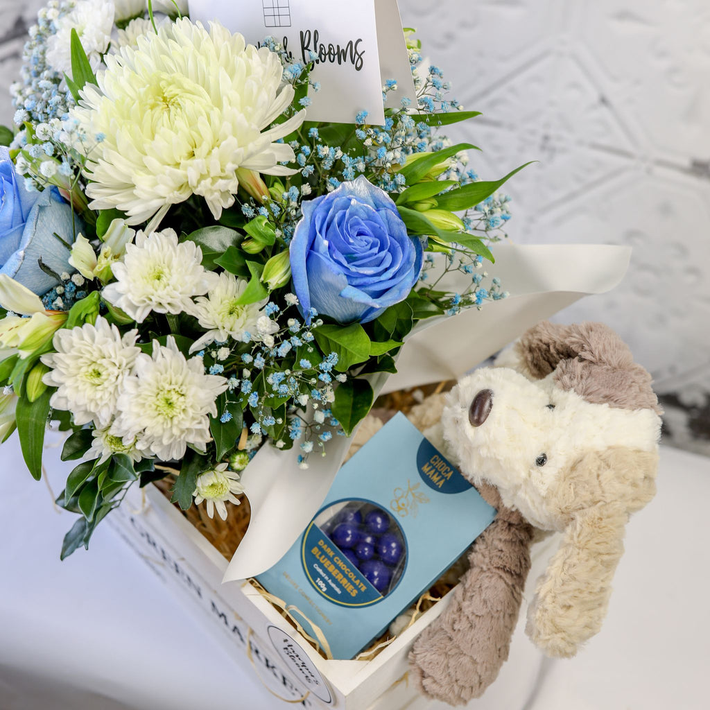 View from above of blue and white flowers, chocolate box and plush puppy arranged in a white crate