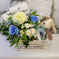 Front view of blue-tinted roses, white blooms, chocolate box and plush puppy in a white timber crate