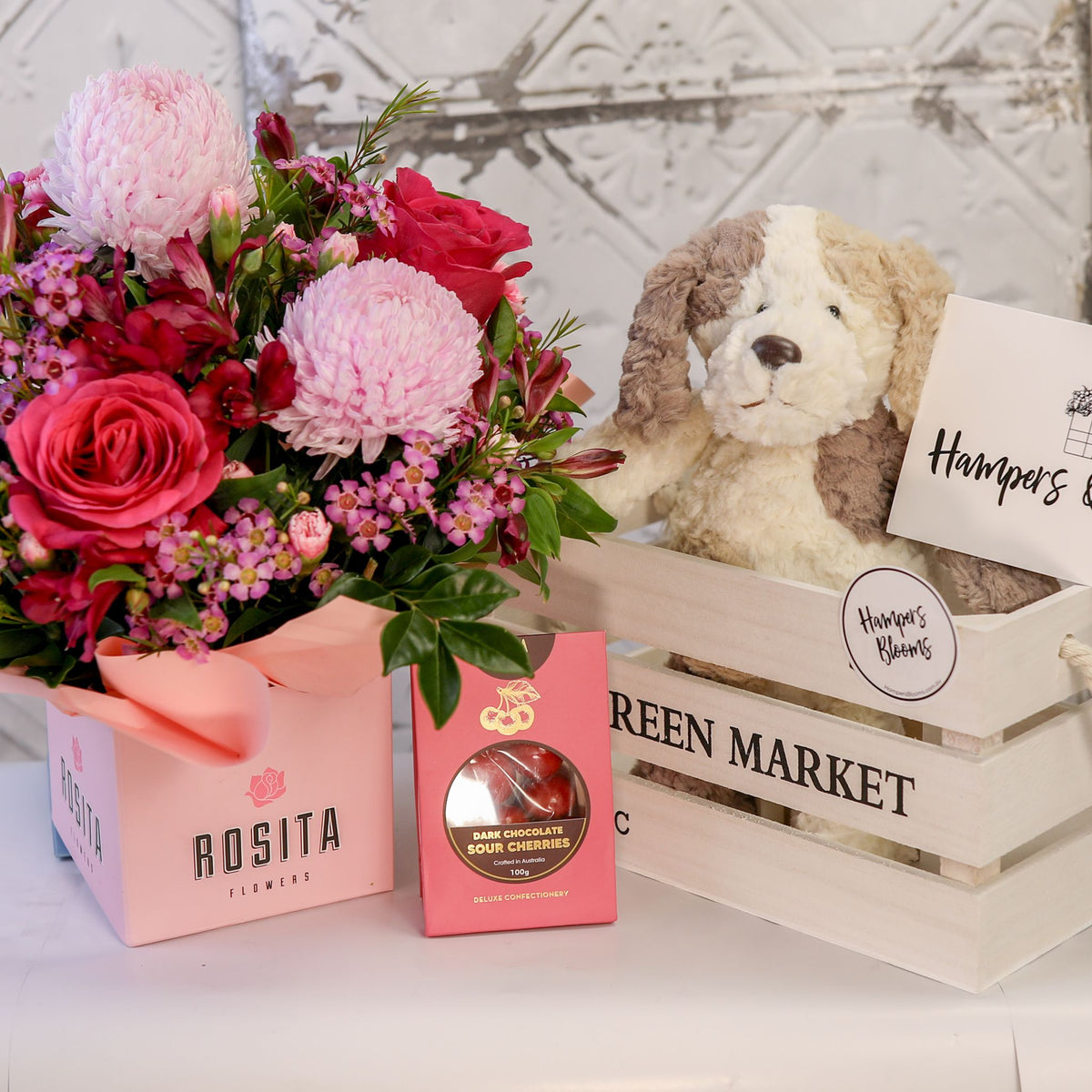 Pink floral pot and chocolate box displayed beside a white crate with a plush puppy and message card