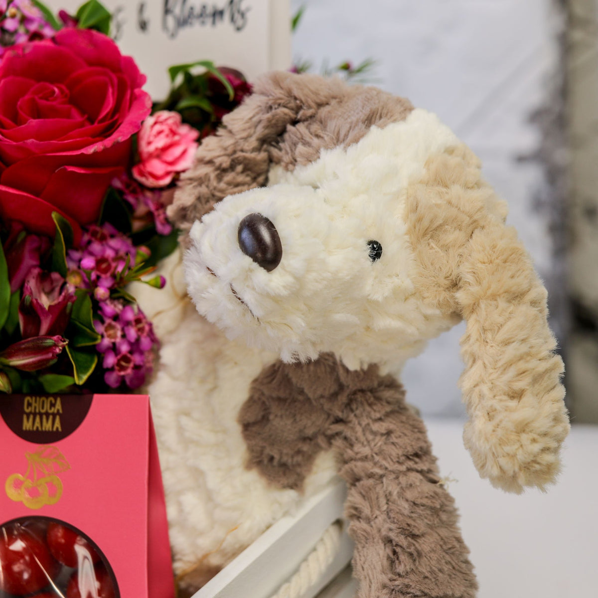 Close-up of pink roses, chocolate box and the plush puppy leaning over the white crate