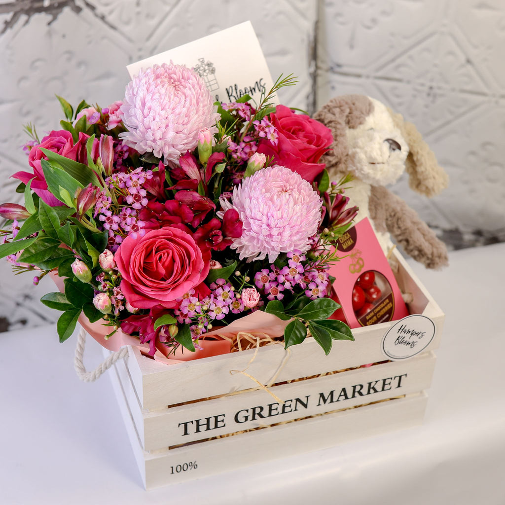 View from above of pink blooms, chocolate box and plush puppy arranged in a white crate