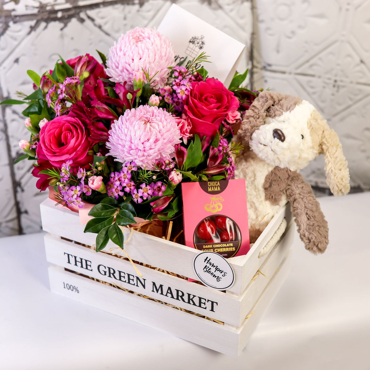 Angled view of pink blooms, chocolate box and plush puppy inside a white timber crate