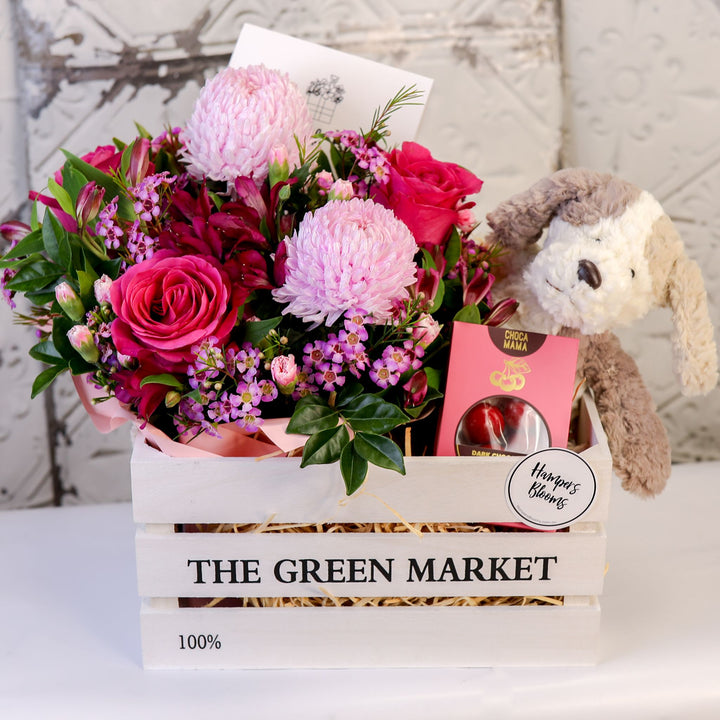 Front view of pink flowers, plush puppy and chocolate box arranged in a white timber crate
