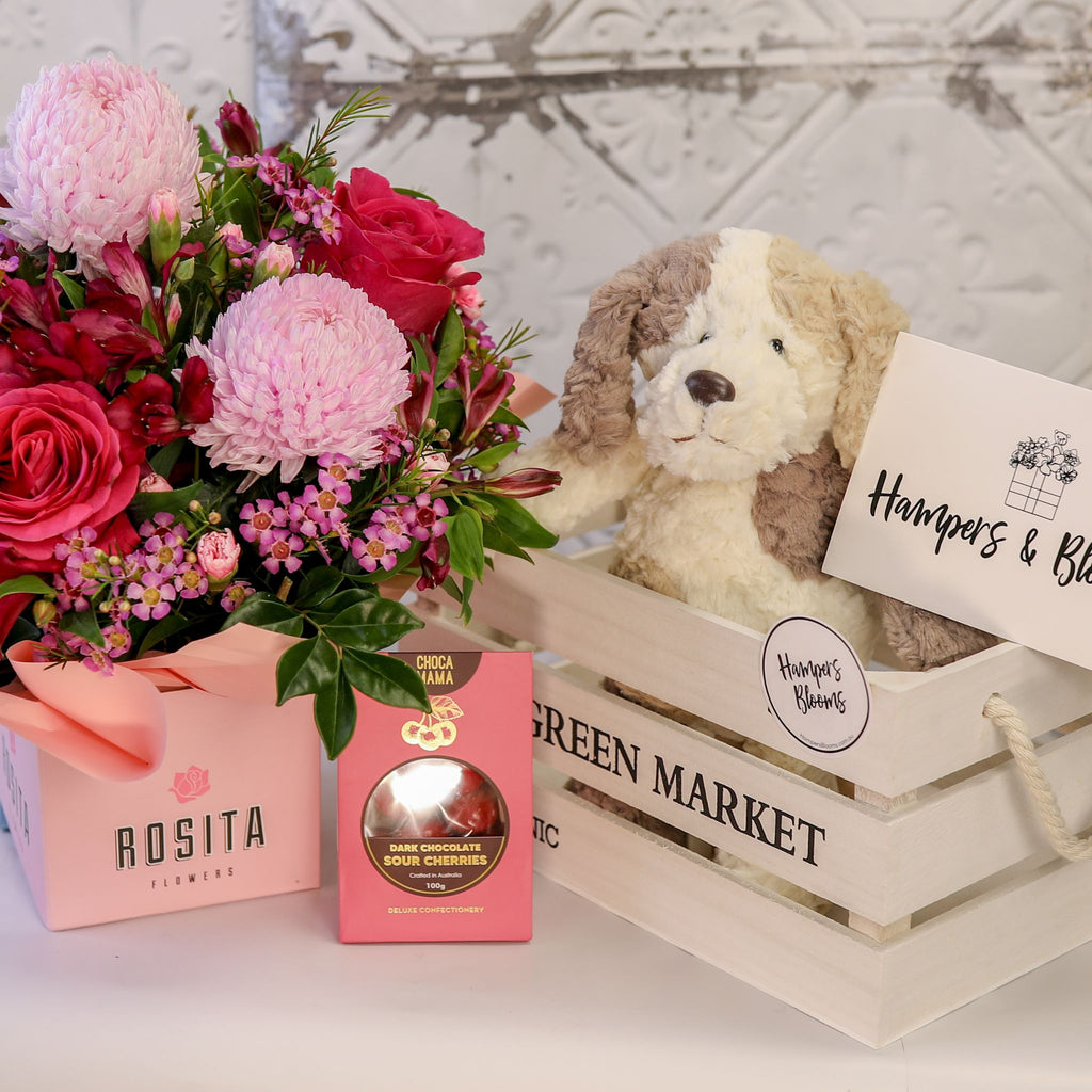 Pink flowers in a ribbon-wrapped pot and chocolate box beside a white crate holding a plush puppy and card