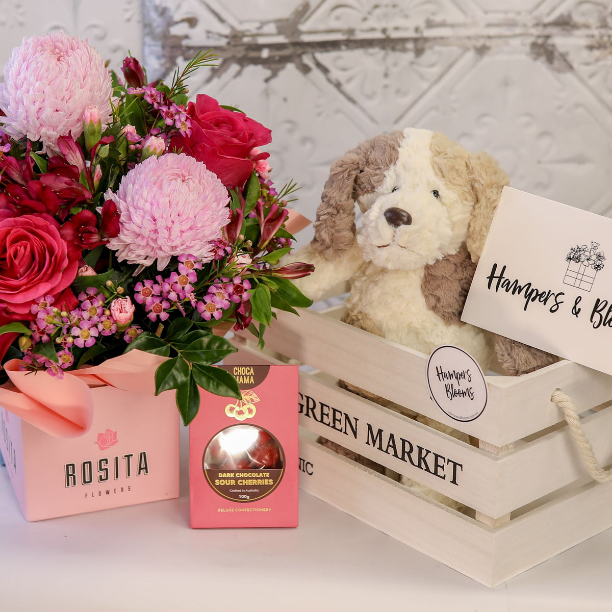 Pink flowers in a ribbon-wrapped pot and chocolate box beside a white crate holding a plush puppy and card