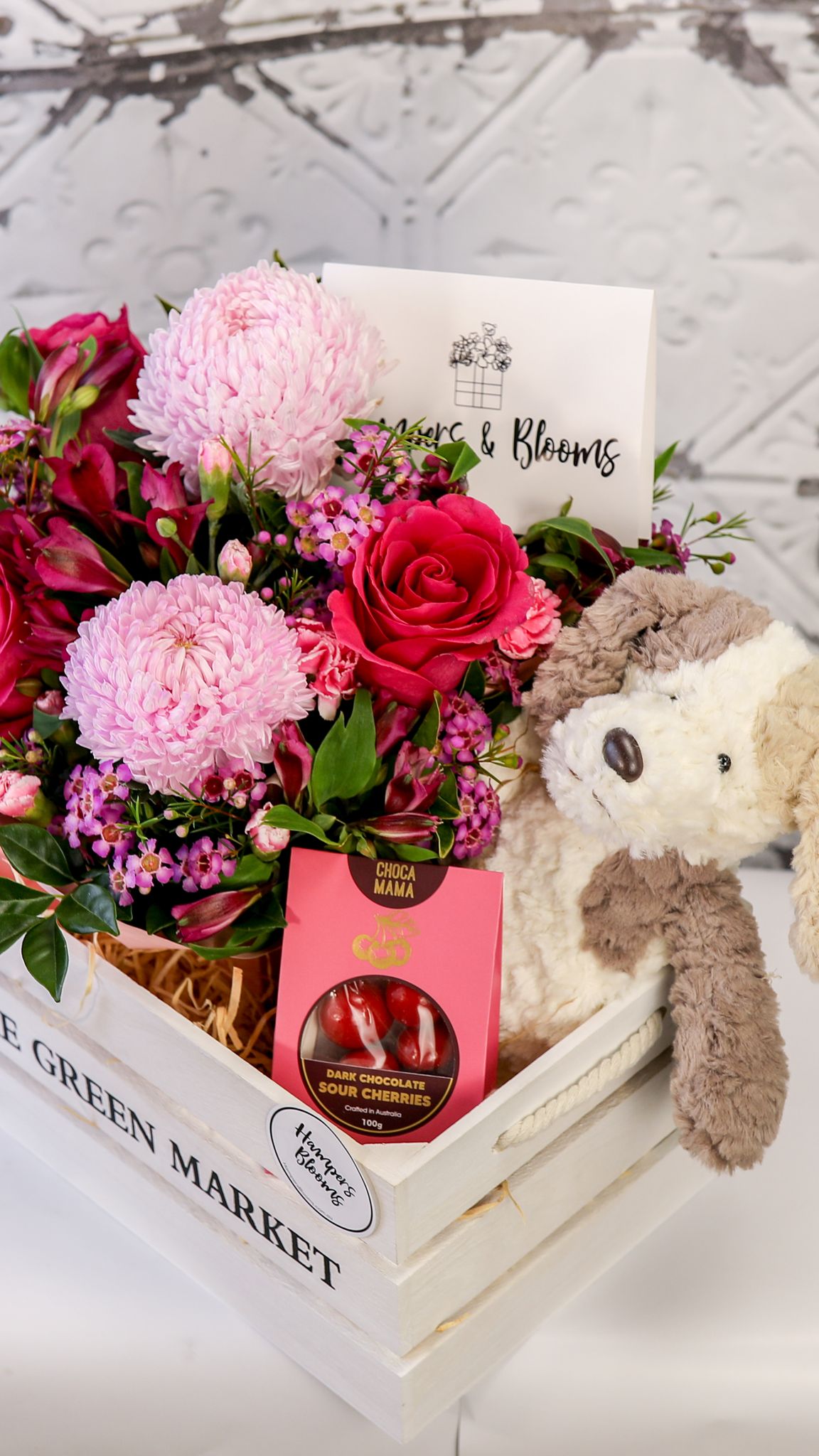 Portrait crop of pink blooms, chocolate box and plush puppy above the corner of a white crate