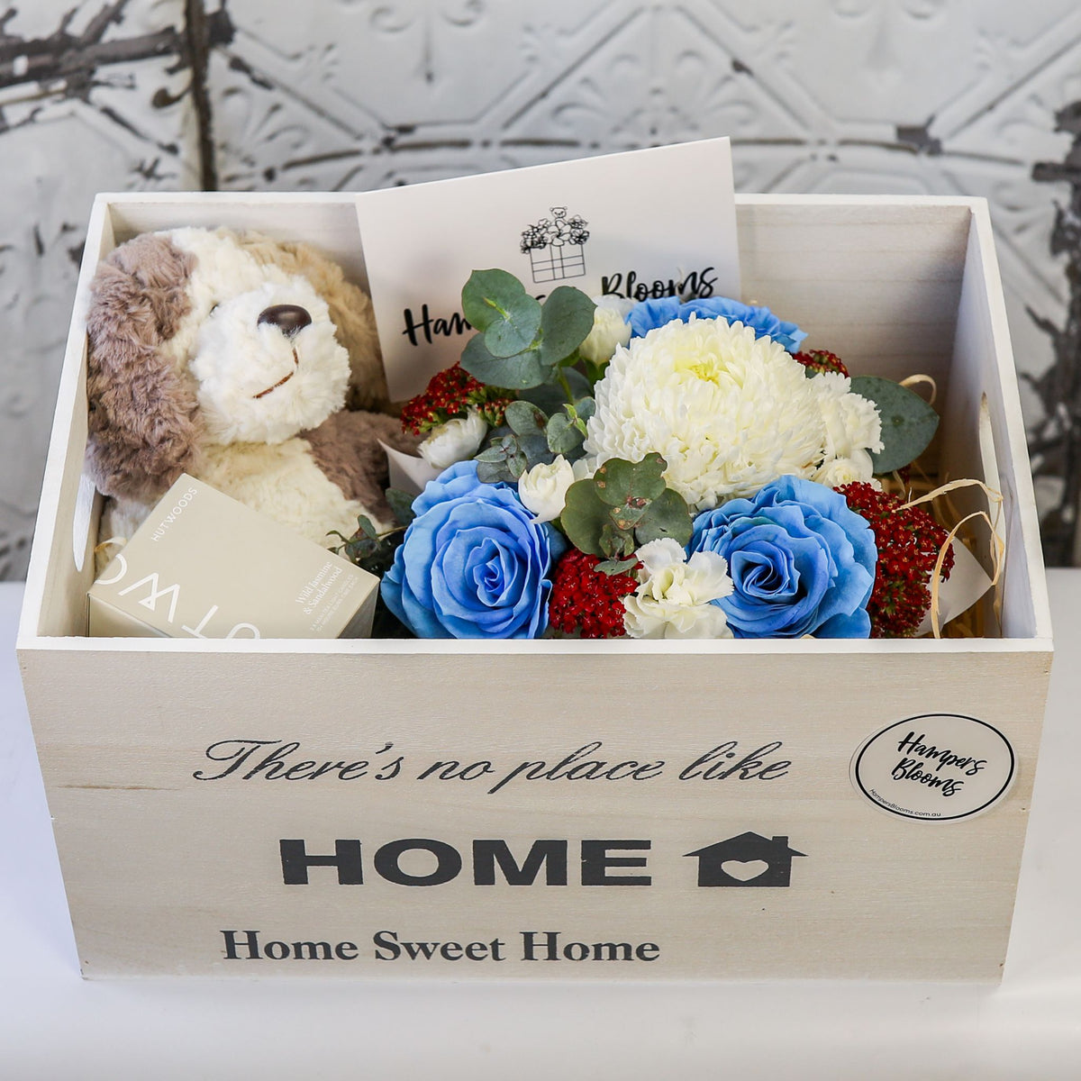 Closer front view of blue and white flowers, plush puppy and boxed candle inside an open white chest