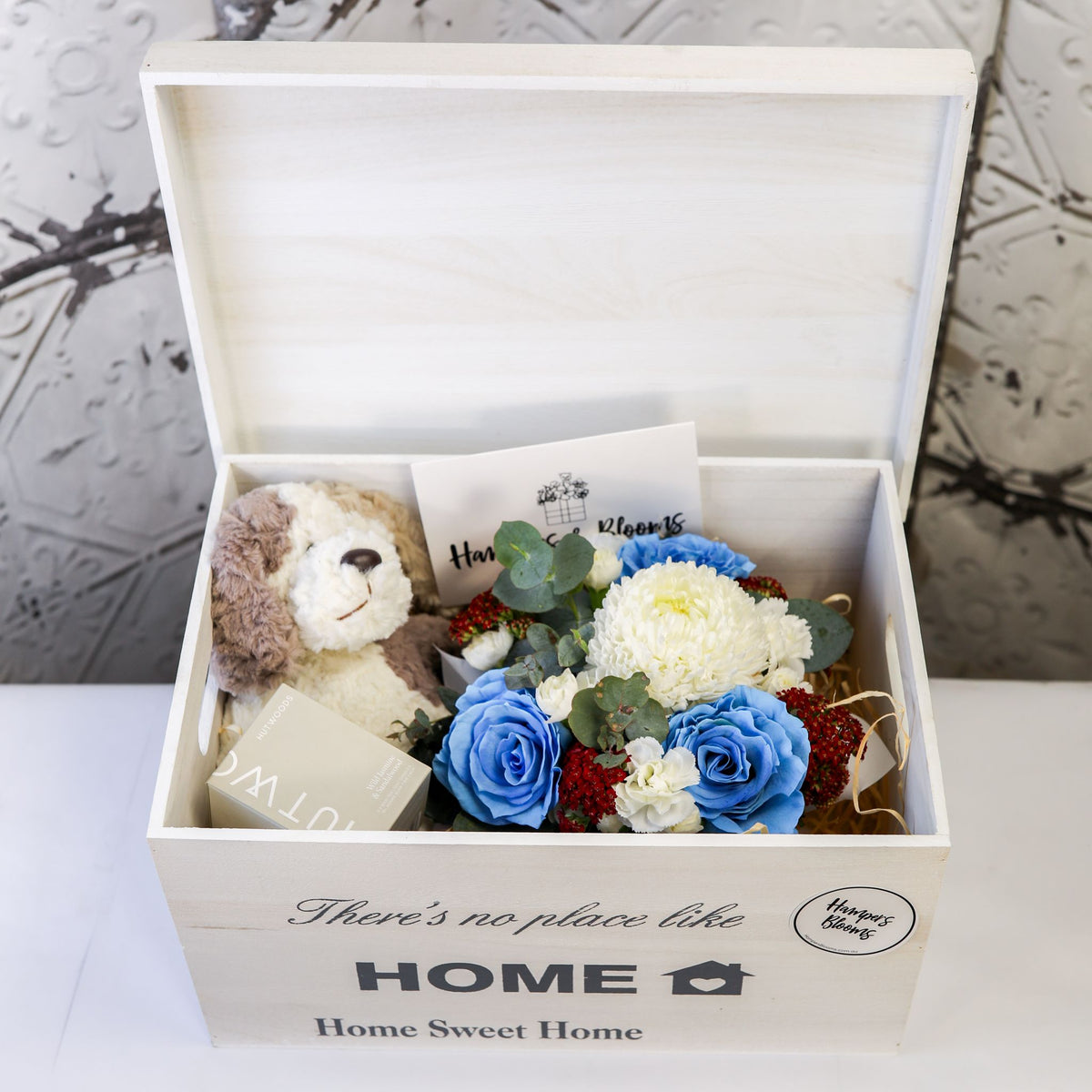 Angled view of an open white chest containing blue and white flowers, plush puppy, candle box and card