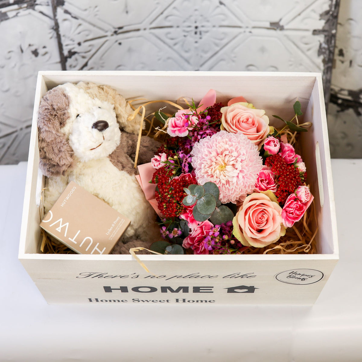 Close-up from above of pink flowers, plush puppy and boxed candle inside a white chest