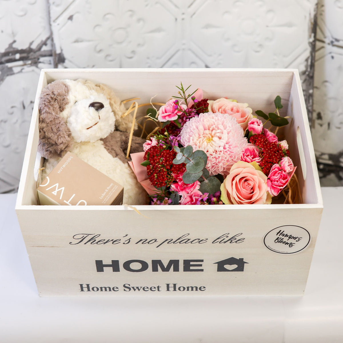 Angled view of pink flowers, plush puppy and boxed candle arranged inside an open white chest