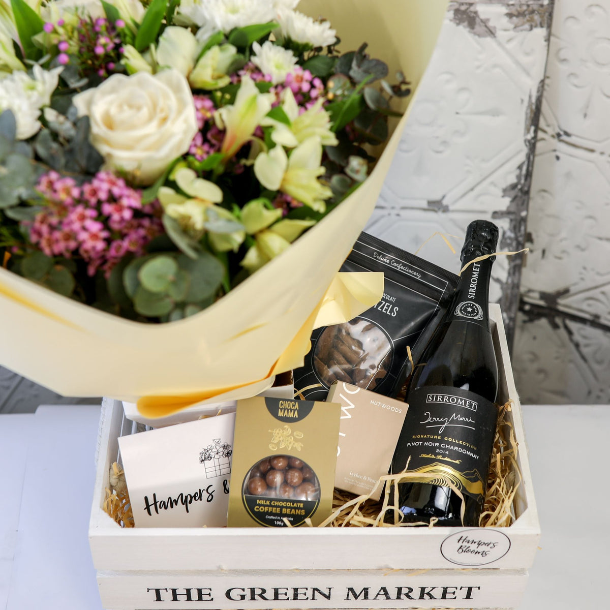 Close-up of white and cream blooms above a bottle, chocolates and candle box inside a white crate