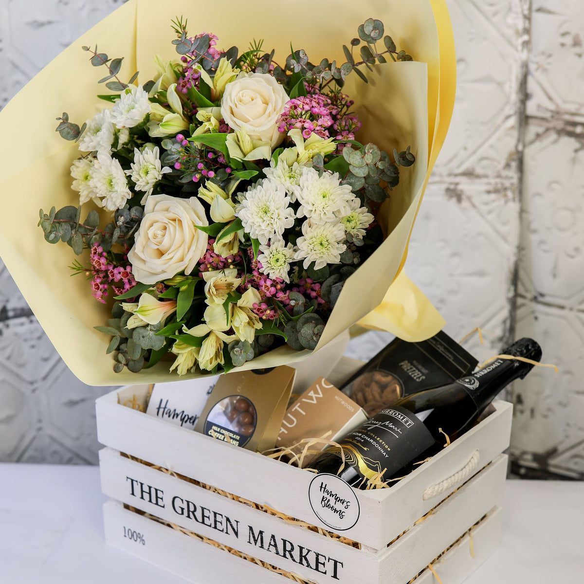 View from above of a white and cream bouquet, bottle and treats arranged in a white crate