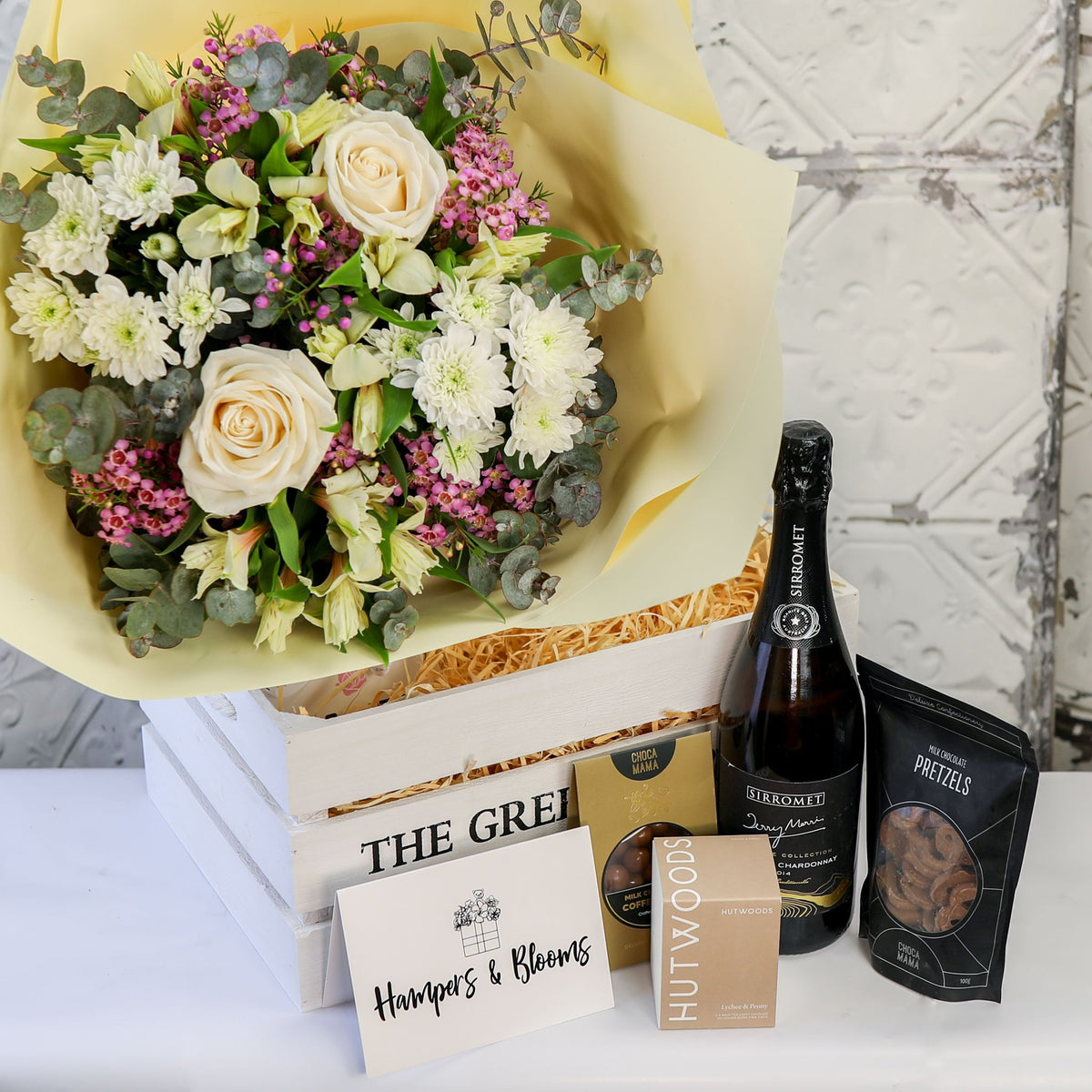 White and cream bouquet above a white crate with bottle, chocolates, candle box and card displayed in front
