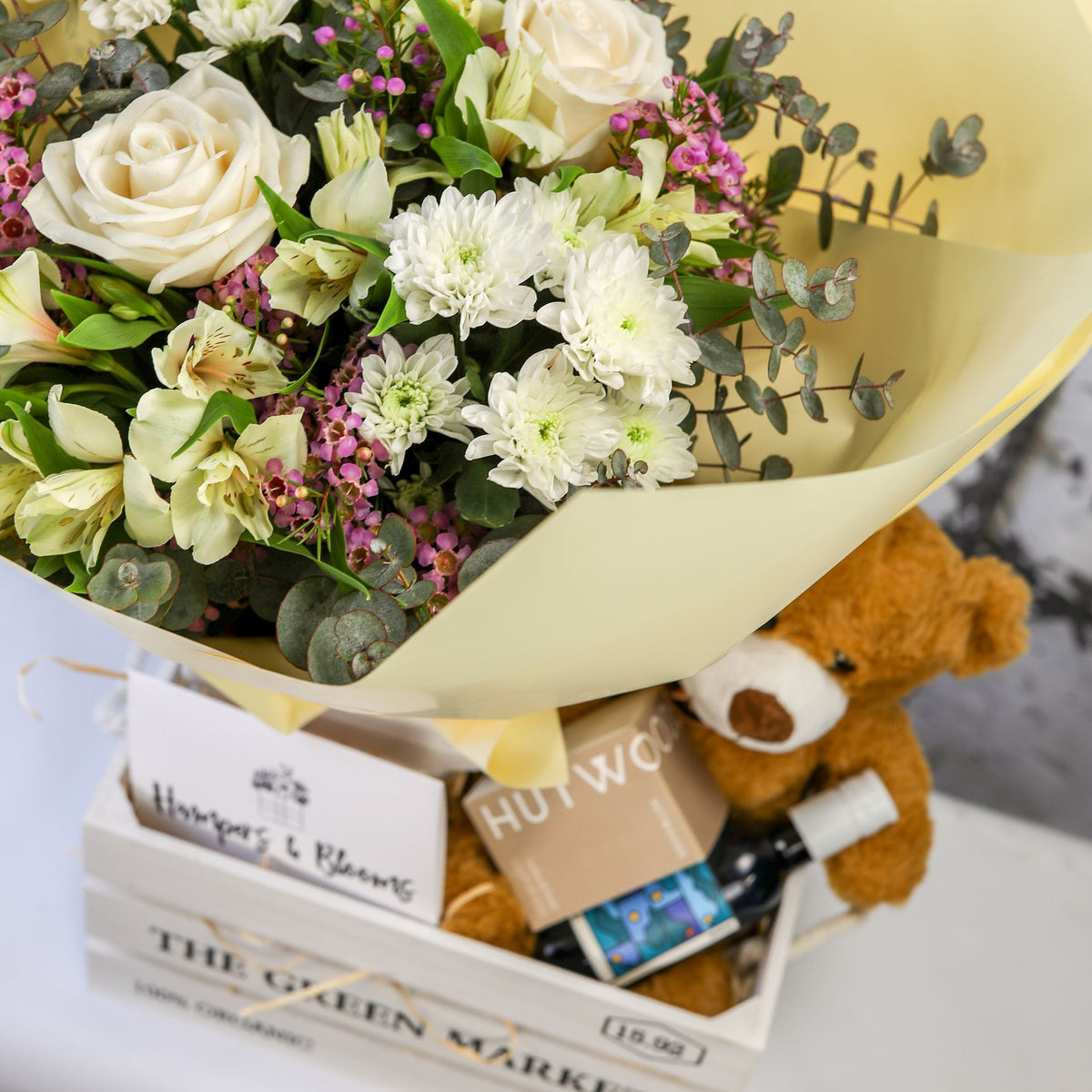 Close-up of white and green blooms above a brown teddy, candle box and small bottle in a white crate