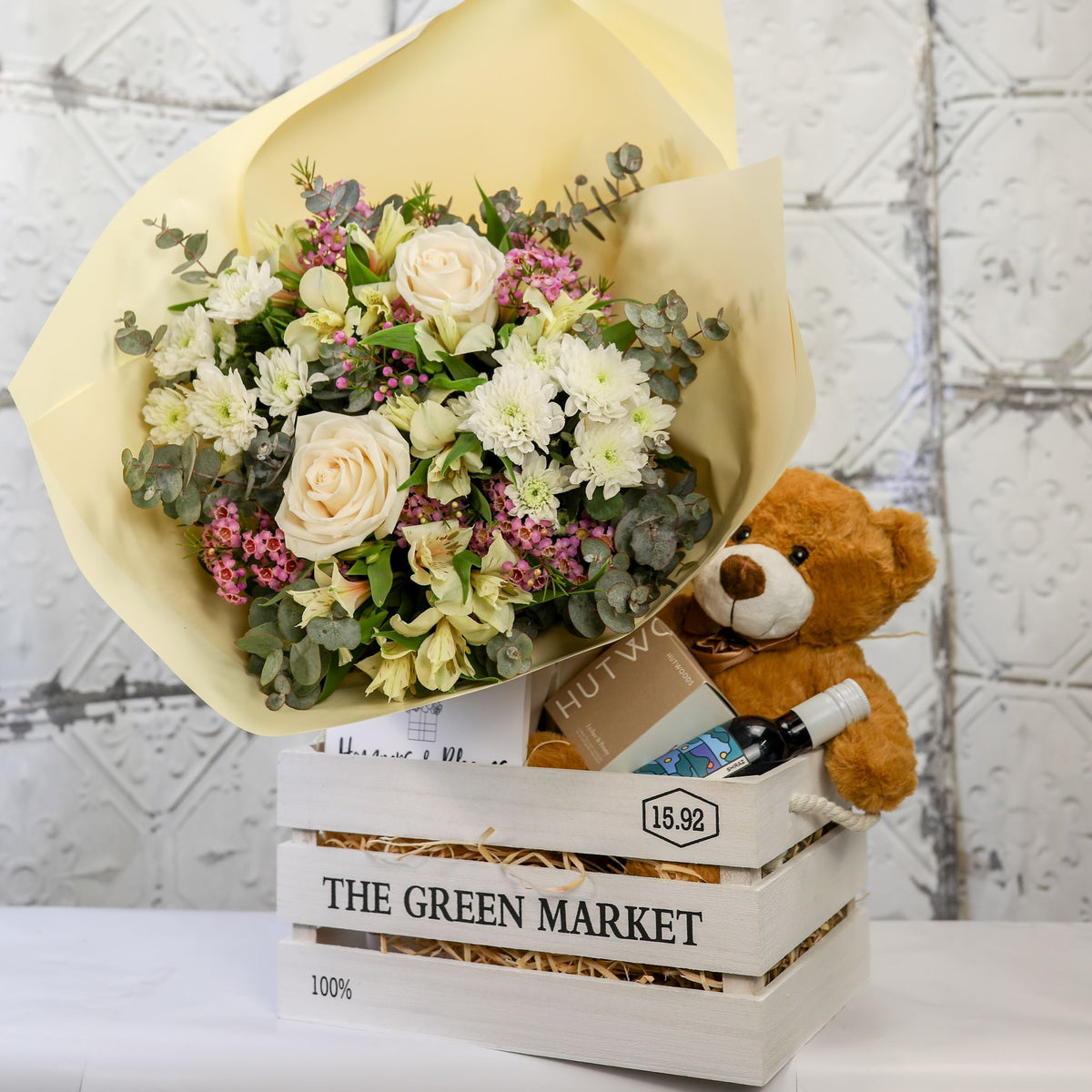 Angled view of a white and green bouquet, brown teddy, candle box and small bottle inside a white crate
