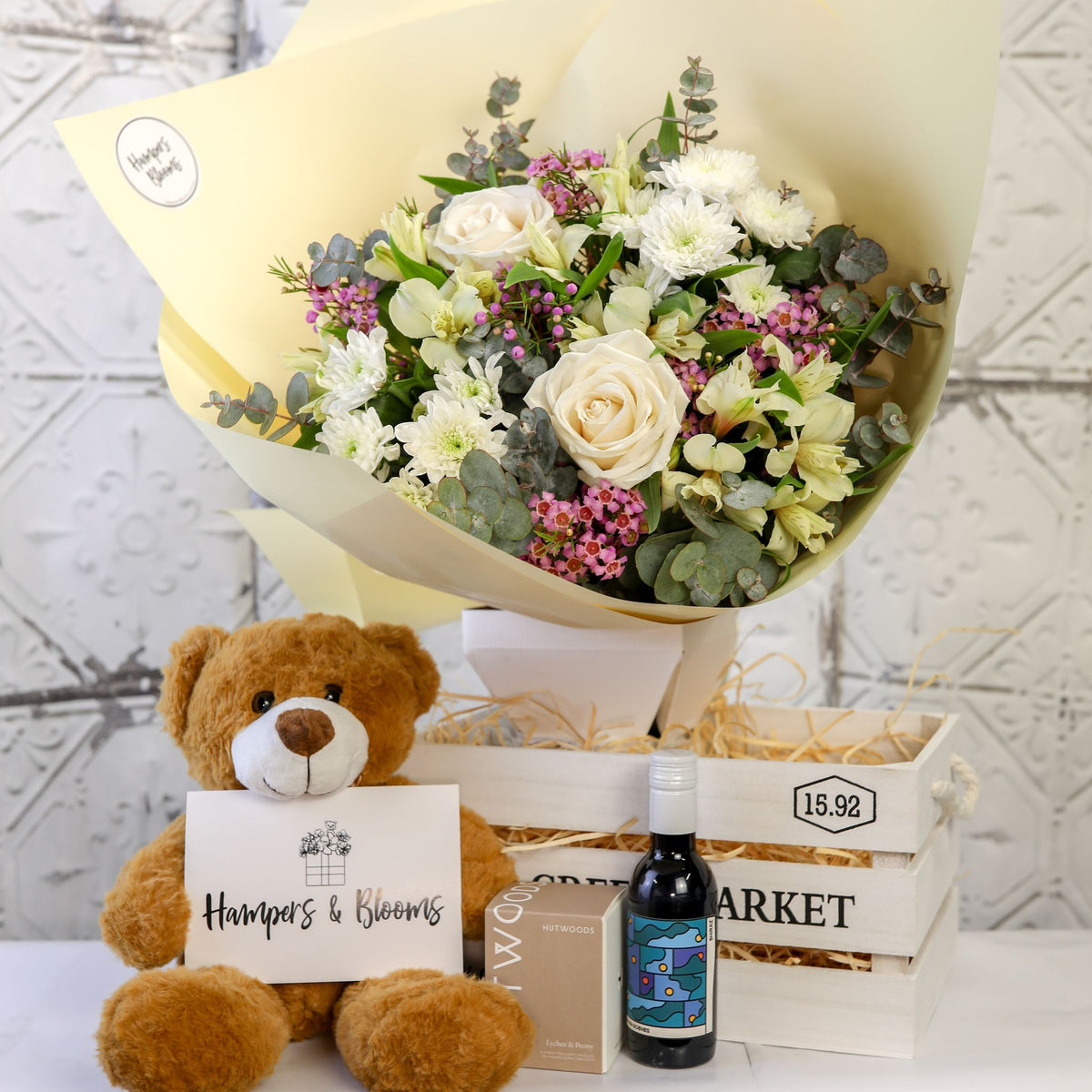 White and green bouquet above a white crate with a seated brown teddy holding a card, candle box and small bottle in front